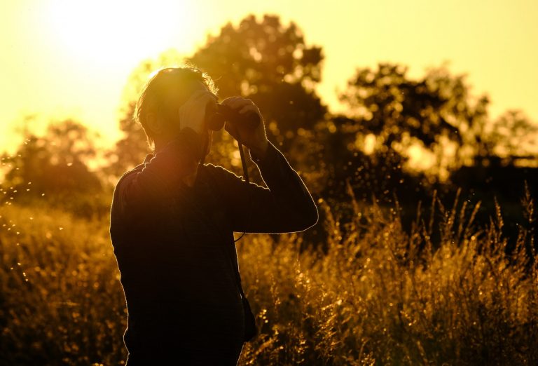 Man looking through his binoculars to see birds in a nearby lake during a fantastic sunset